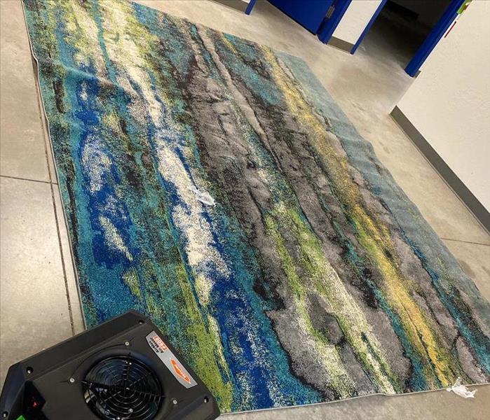 Blue, green, yellow, and gray rug on a gray concrete floor with a black commercial dryer to the left of it. 