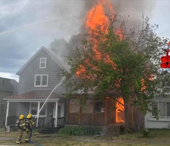 Gray wooden house on fire with firefighters working in the front lawn.